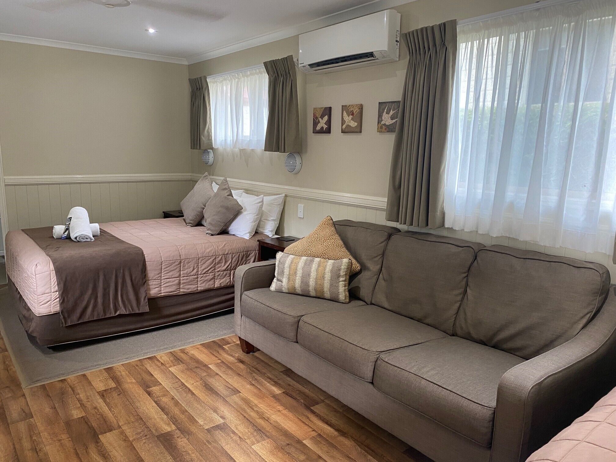 Una habitación de hotel con cama, sofá, suelo de madera, unidad de aire acondicionado y cortinas.
