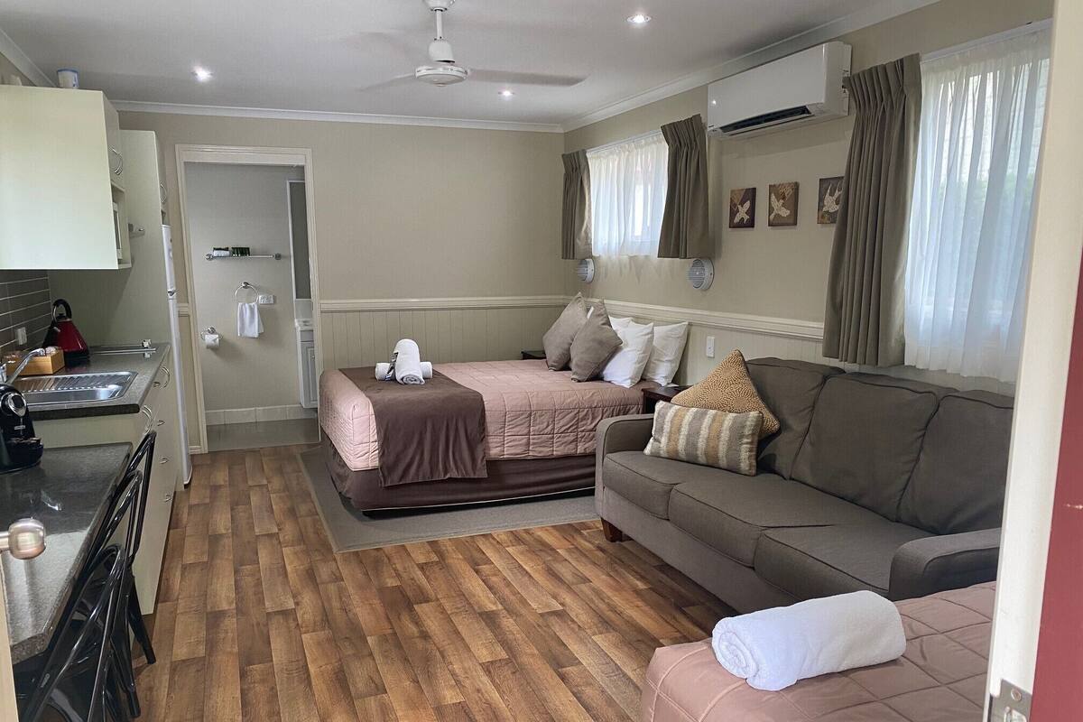 Habitación de hotel compacta con kitchenette, una cama con dos almohadas, un sofá y una ventana con cortinas.