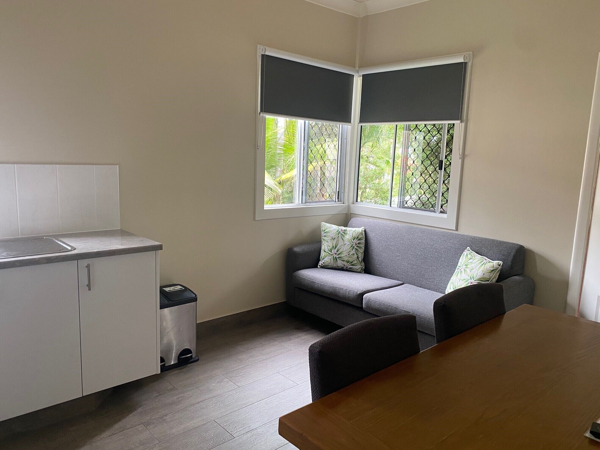 A compact kitchen with a sink, cabinets, and a trash bin. There is a grey sofa with patterned cushions and a wooden dining table.