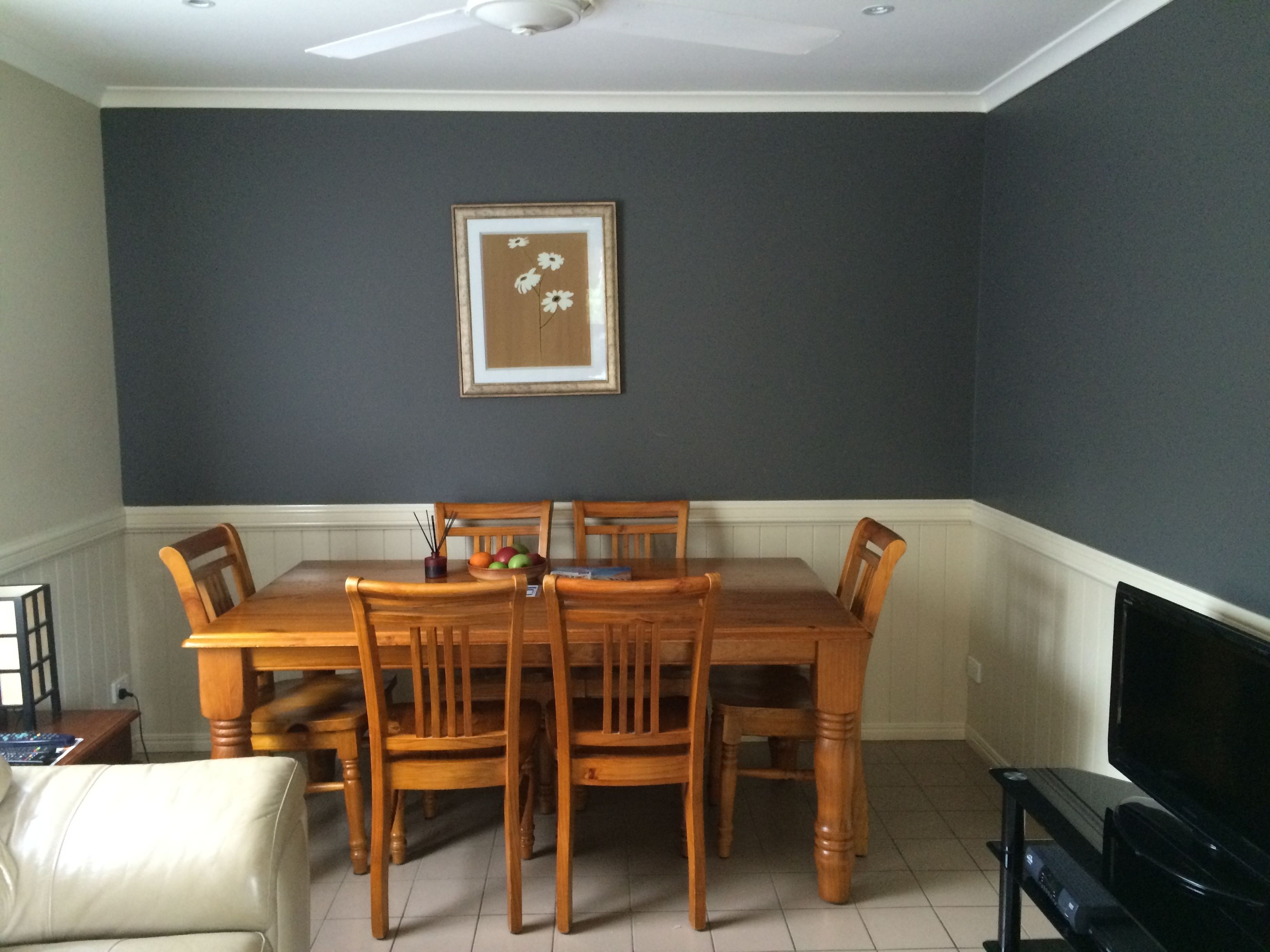 Uma sala de jantar com mesa e cadeiras de madeira, um quadro pendurado na parede e uma televisão sobre um móvel.
