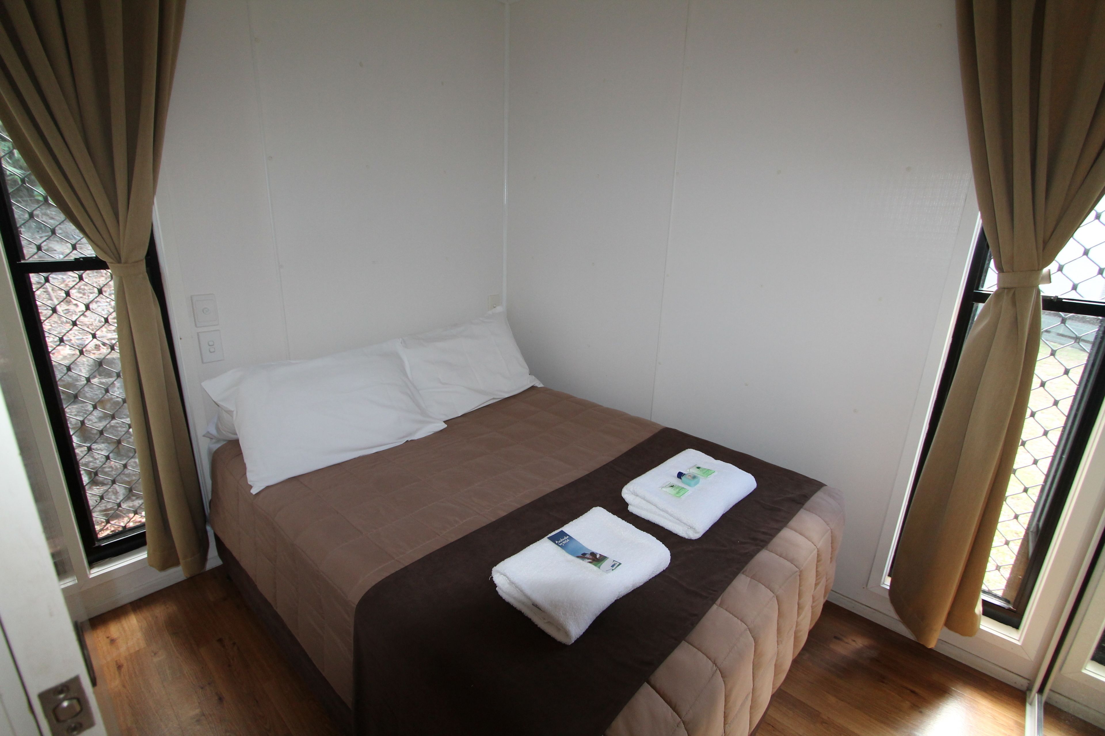 A single bed with a brown bedspread and white pillows, two folded towels on the bed, and a window with curtains.