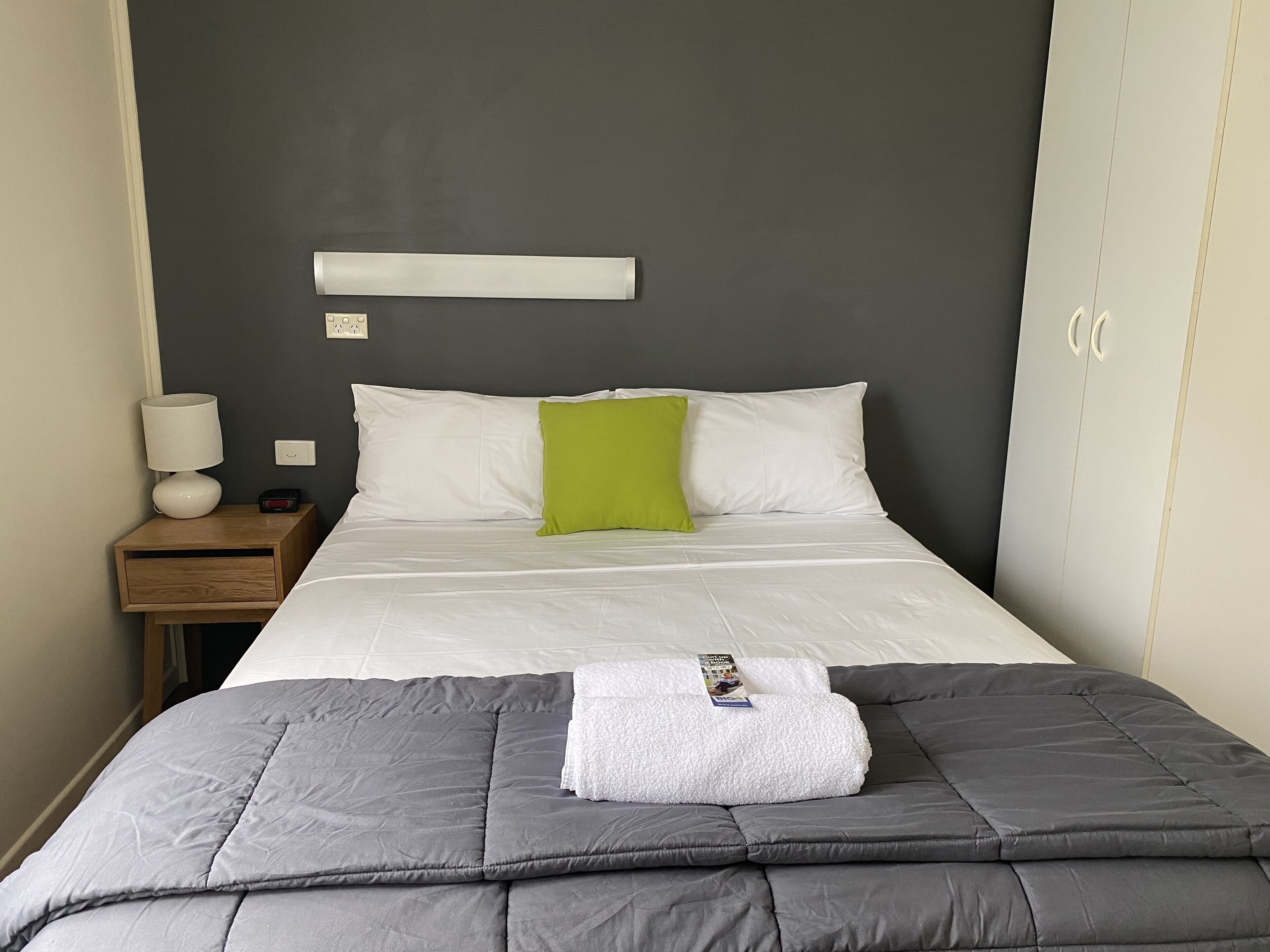 A neatly made bed with a grey duvet, a white pillow, and a green accent pillow. There is a white lamp on a wooden nightstand beside the bed.