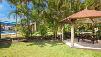 A covered outdoor area with a picnic table, surrounded by palm trees and a pool.
