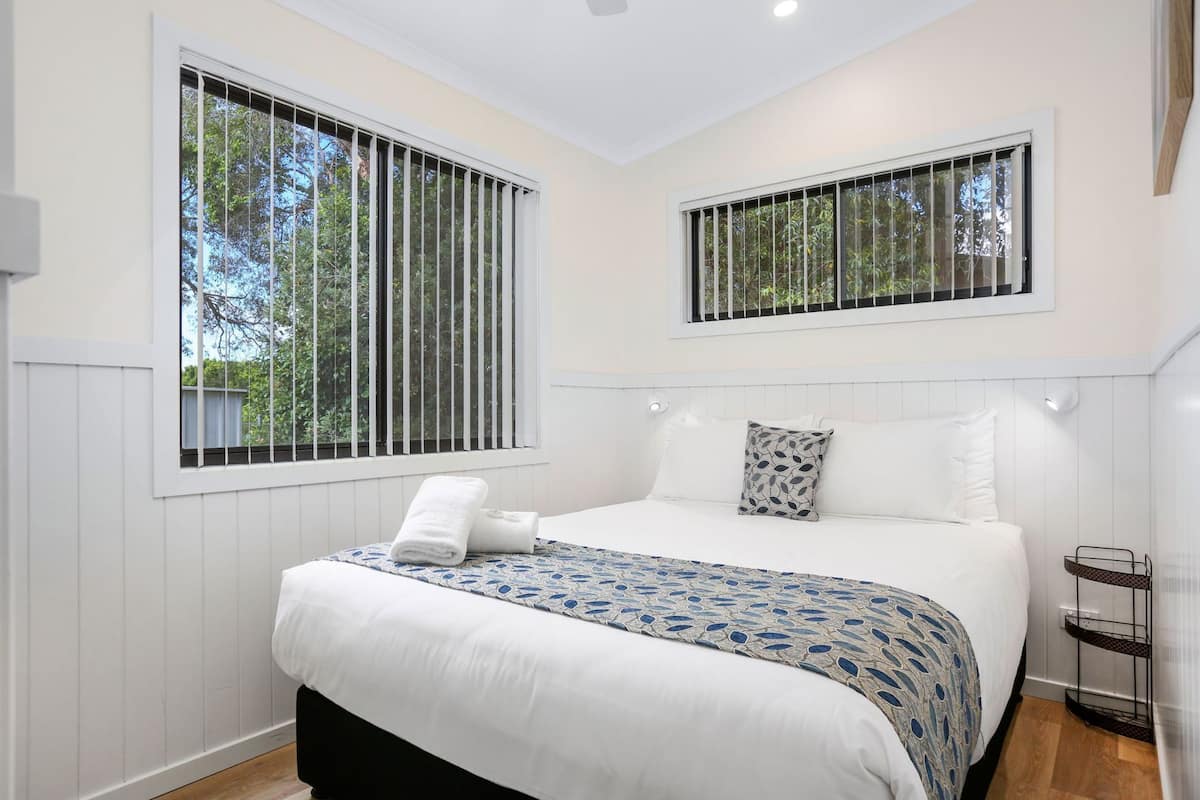 A bedroom with a bed, two windows with blinds, a bedside table, and a nightstand.