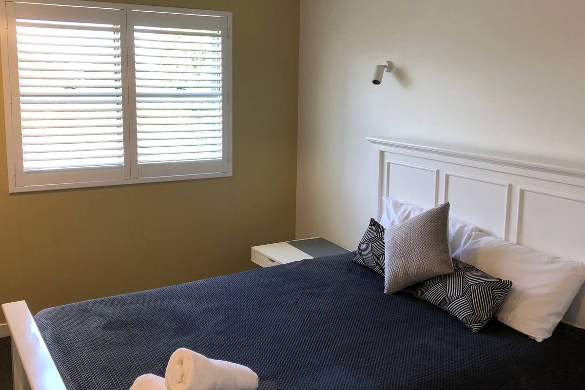 Una cama bien hecha con una colcha azul oscuro y almohadas blancas, una ventana con persianas y una mesita de noche blanca.