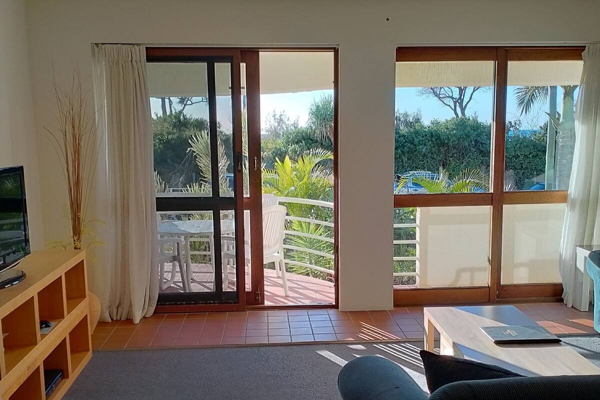 Una sala de estar con un sofá, una mesa de centro, un televisor en un estante de madera y un ventanal corredizo que da a un balcón con muebles de exterior.