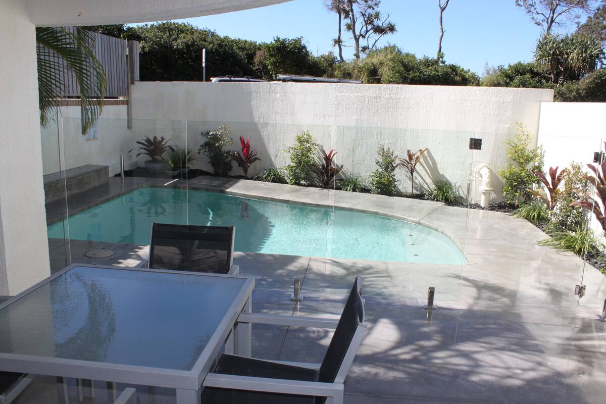 Zona de piscina exterior moderna con una mesa y sillas de vidrio, rodeada de plantas variadas y un muro blanco.