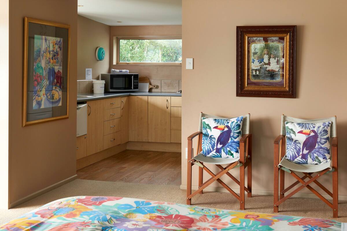 A bedroom with a bed, two chairs with tropical prints, a kitchen with wooden cabinets, a microwave, and a window with a view of greenery.