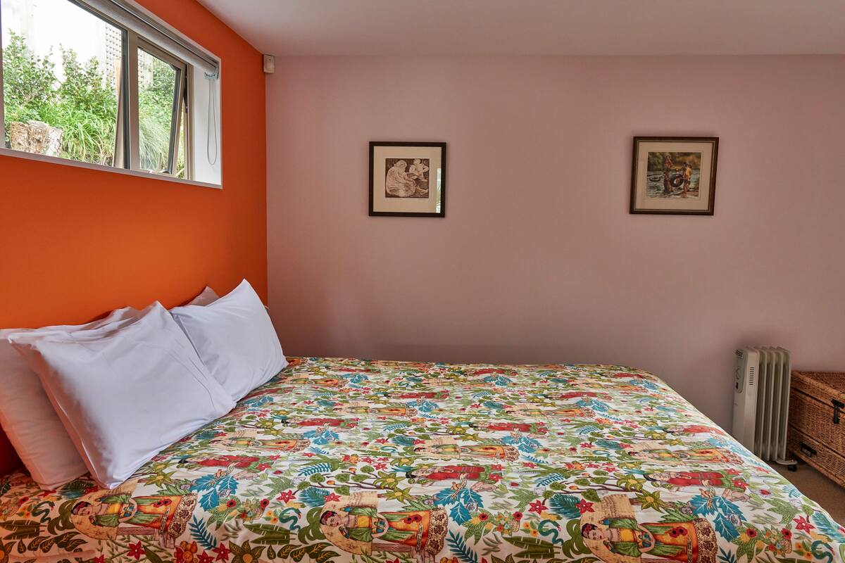 A bedroom with an orange wall, a bed with white pillows, a colorful bedspread, two framed pictures on the wall, and a radiator.