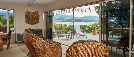 A spacious living area with wicker furniture, a large sliding glass door leading to an outdoor deck with a view of mountains and a lake.