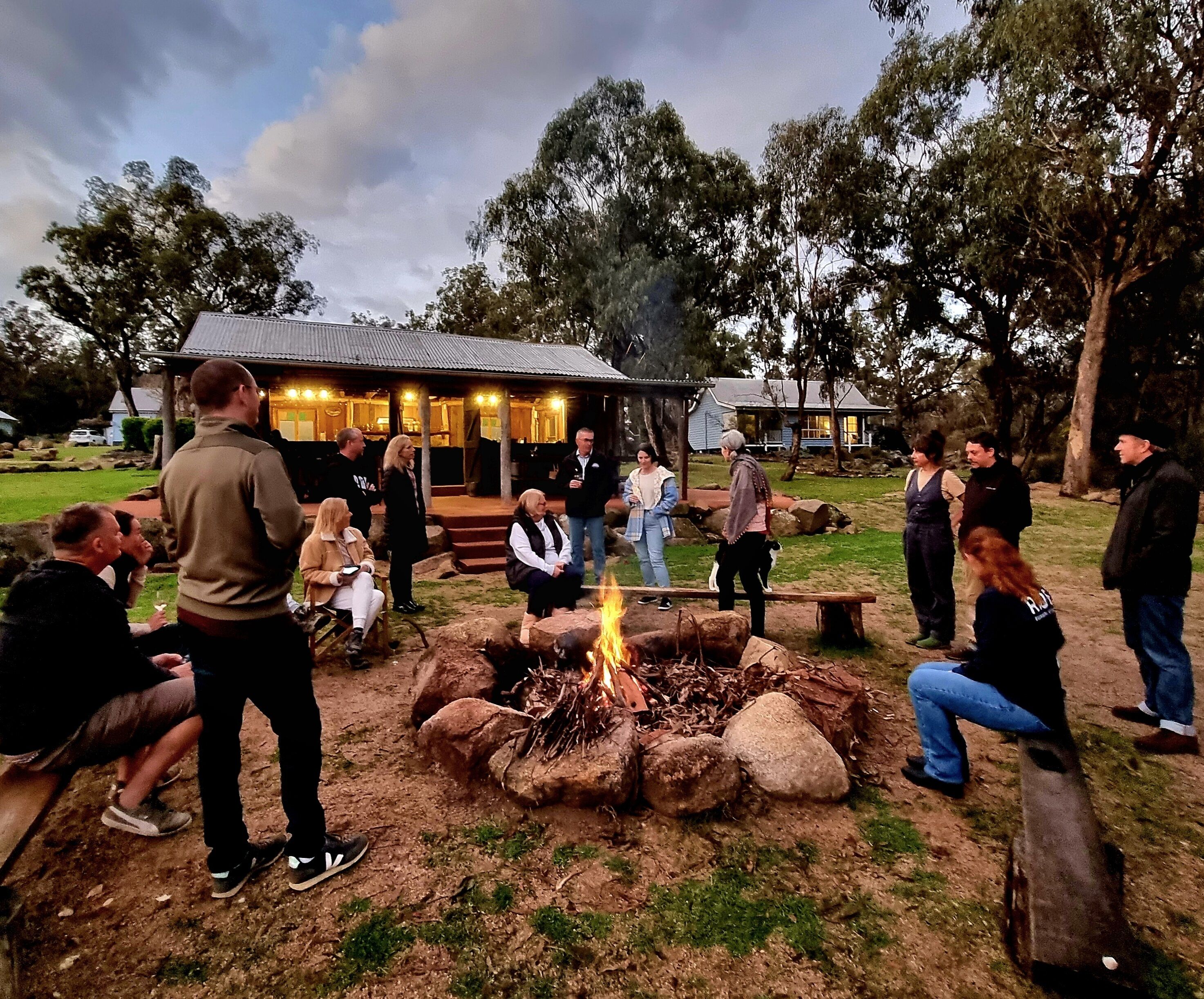 Un grupo de personas reunidas alrededor de un hogar en un espacio al aire libre, con un edificio al fondo.