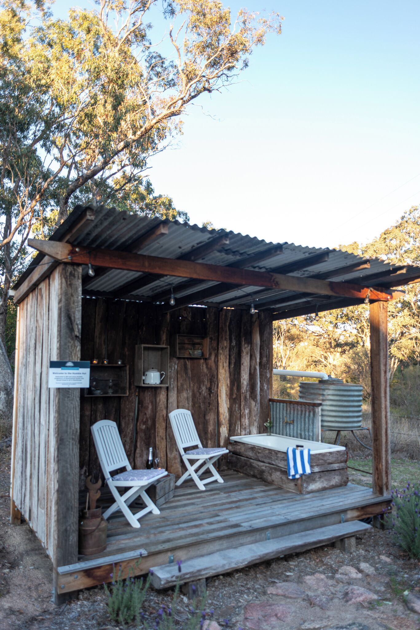A rustic wooden cabin with two white chairs and a bench, a small table, and a metal water tank.