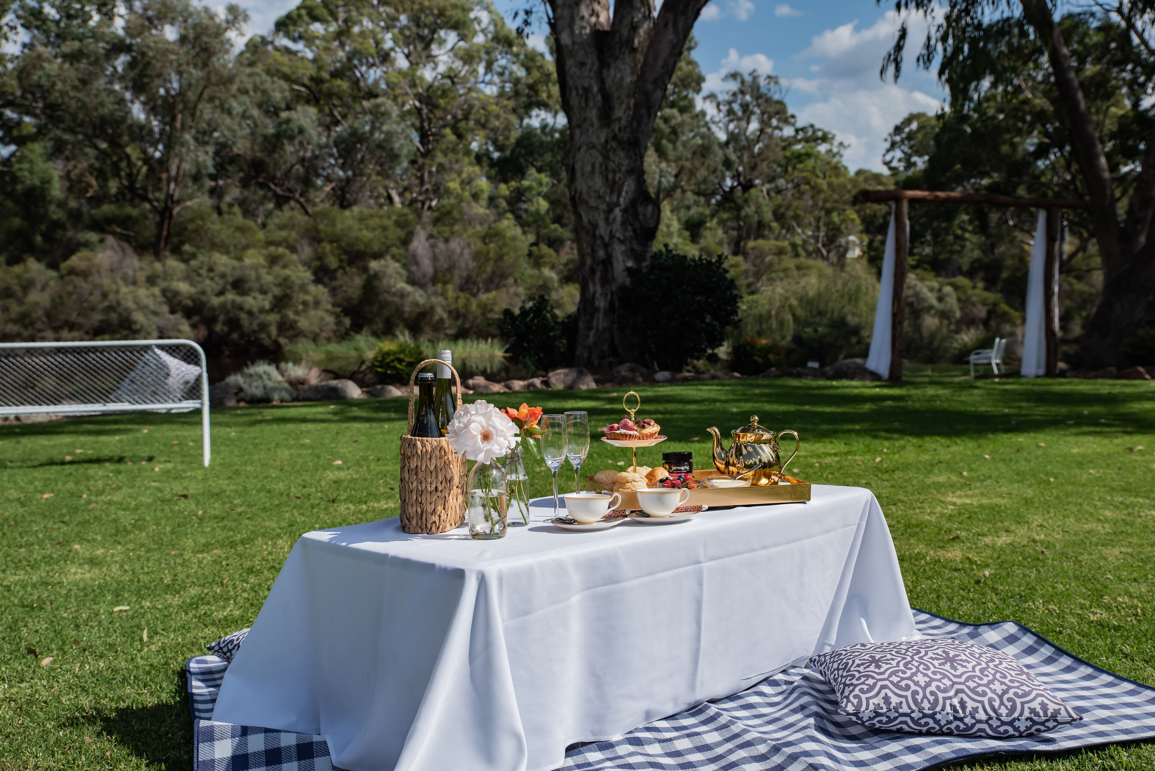 An outdoor dining setup with a table, picnic blanket, and food items.