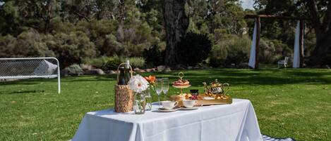 An outdoor dining setup with a table, picnic blanket, and food items.