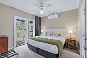 Un dormitorio con una cama grande, un ventilador de techo, una mesita de noche con lámpara y vistas a un área exterior a través de una puerta corrediza de vidrio.