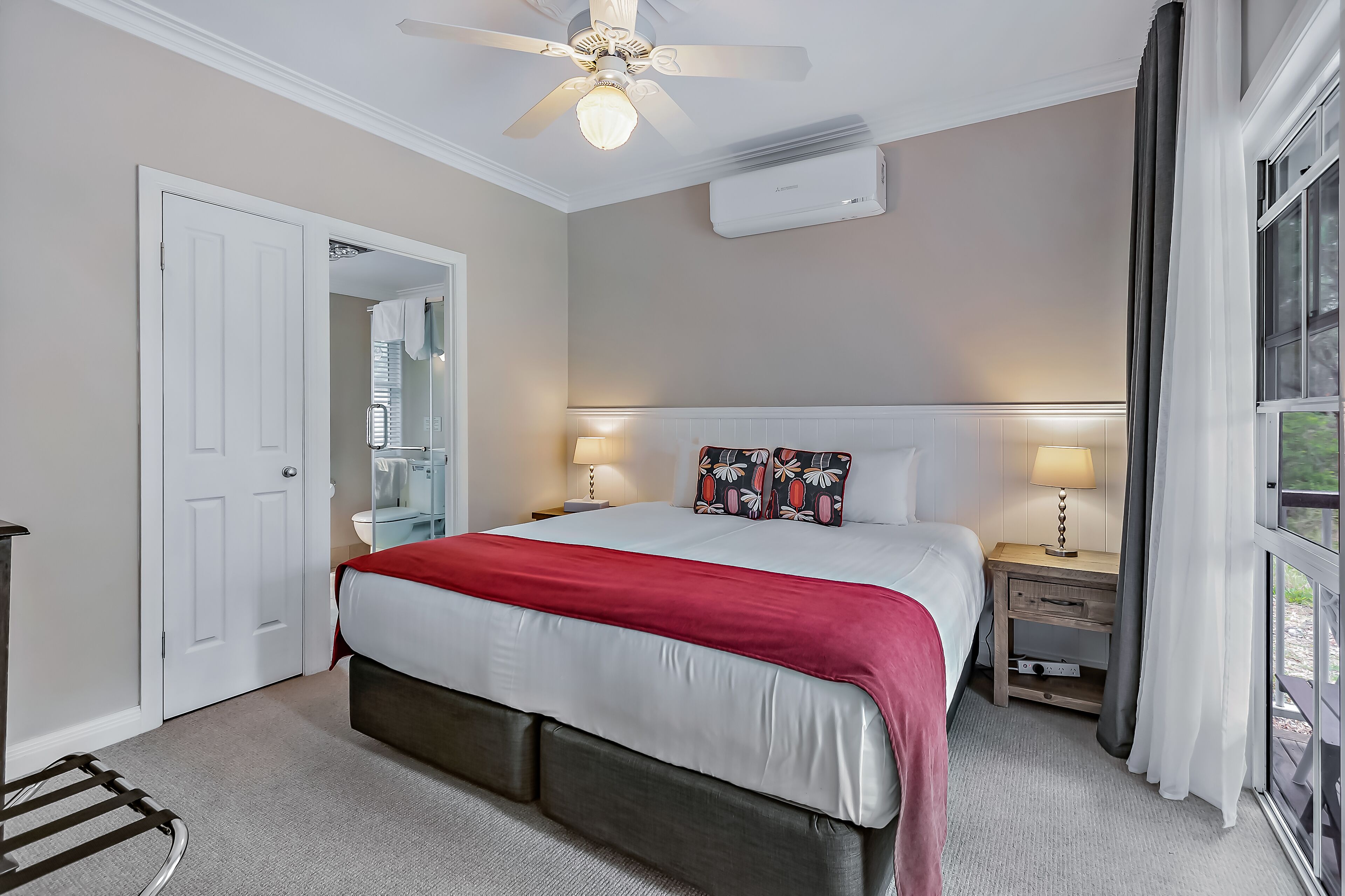 A hotel room with a bed, bedside tables, a ceiling fan, an air conditioning unit, and a window with curtains.
