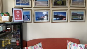 Une chambre d’hôtel avec un mur recouvert de photographies de paysages encadrées, un mini-bar contenant des collations et des boissons, et un canapé rouge agrémenté de coussins à motifs.