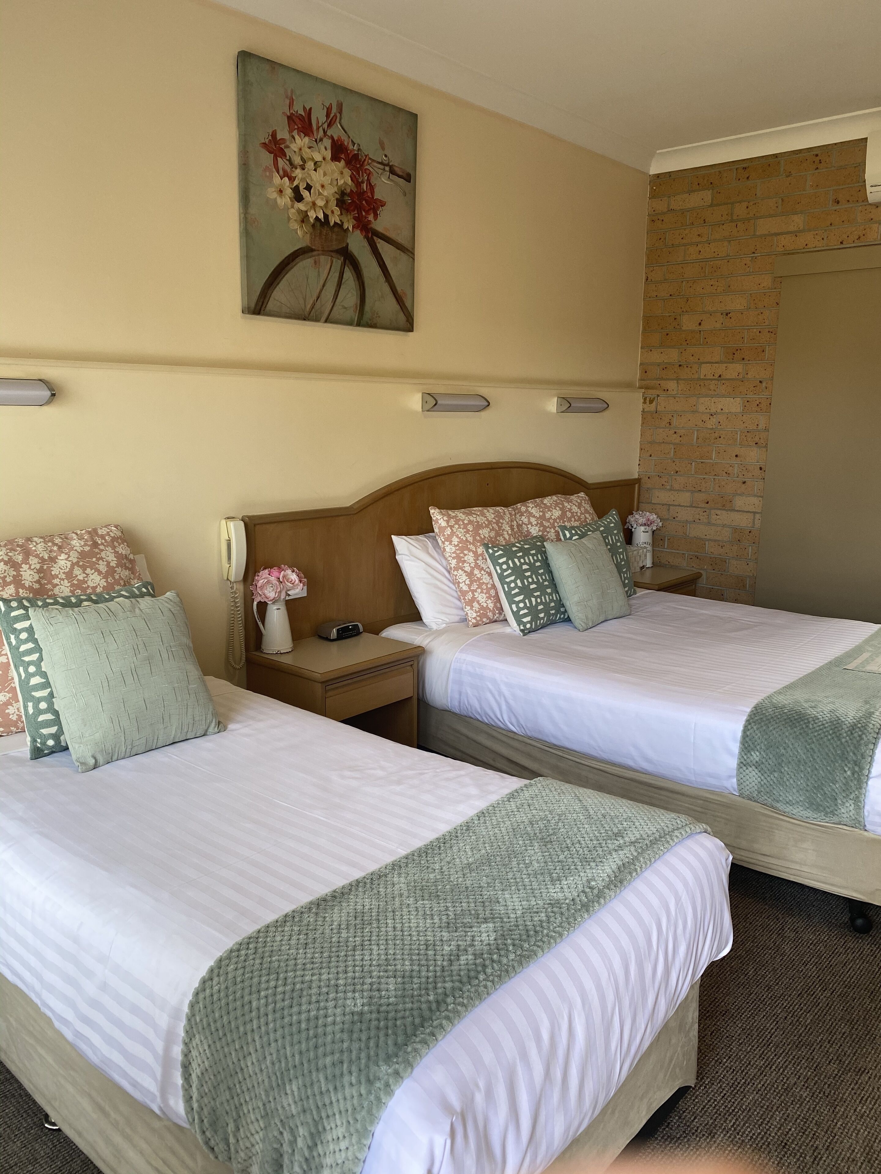 A hotel room with two single beds, a wooden headboard, a nightstand with a phone and a lamp, a painting of flowers on the wall, and a brick wall.