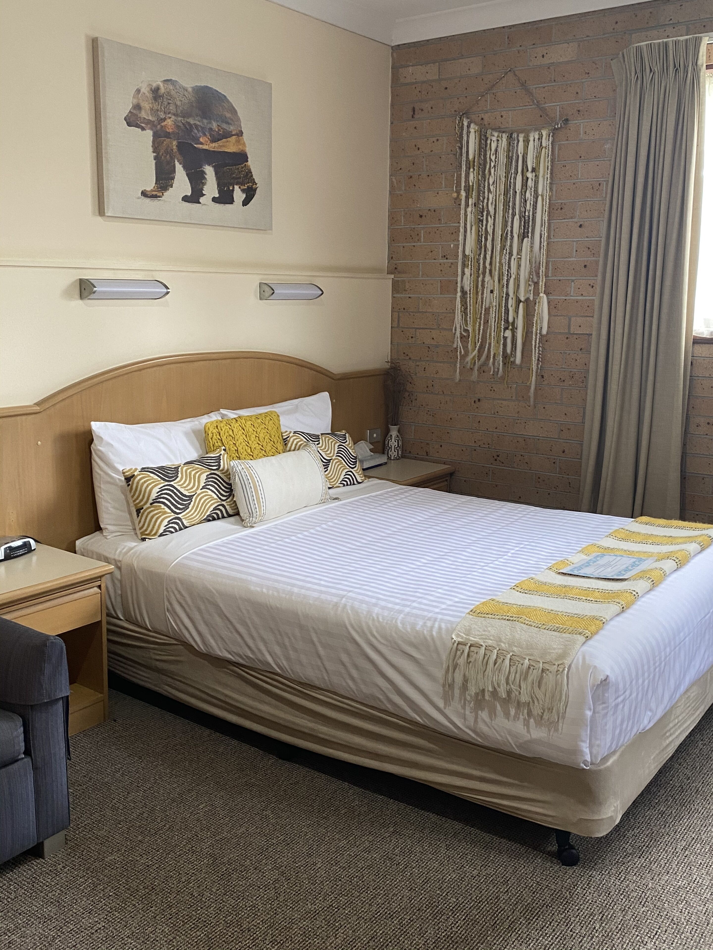 Hotelkamer met een groot bed, een hoofdbord, nachtkastje, lamp, een wanddecoratie van een beer, een dreamcatcher en een raam met gordijnen.