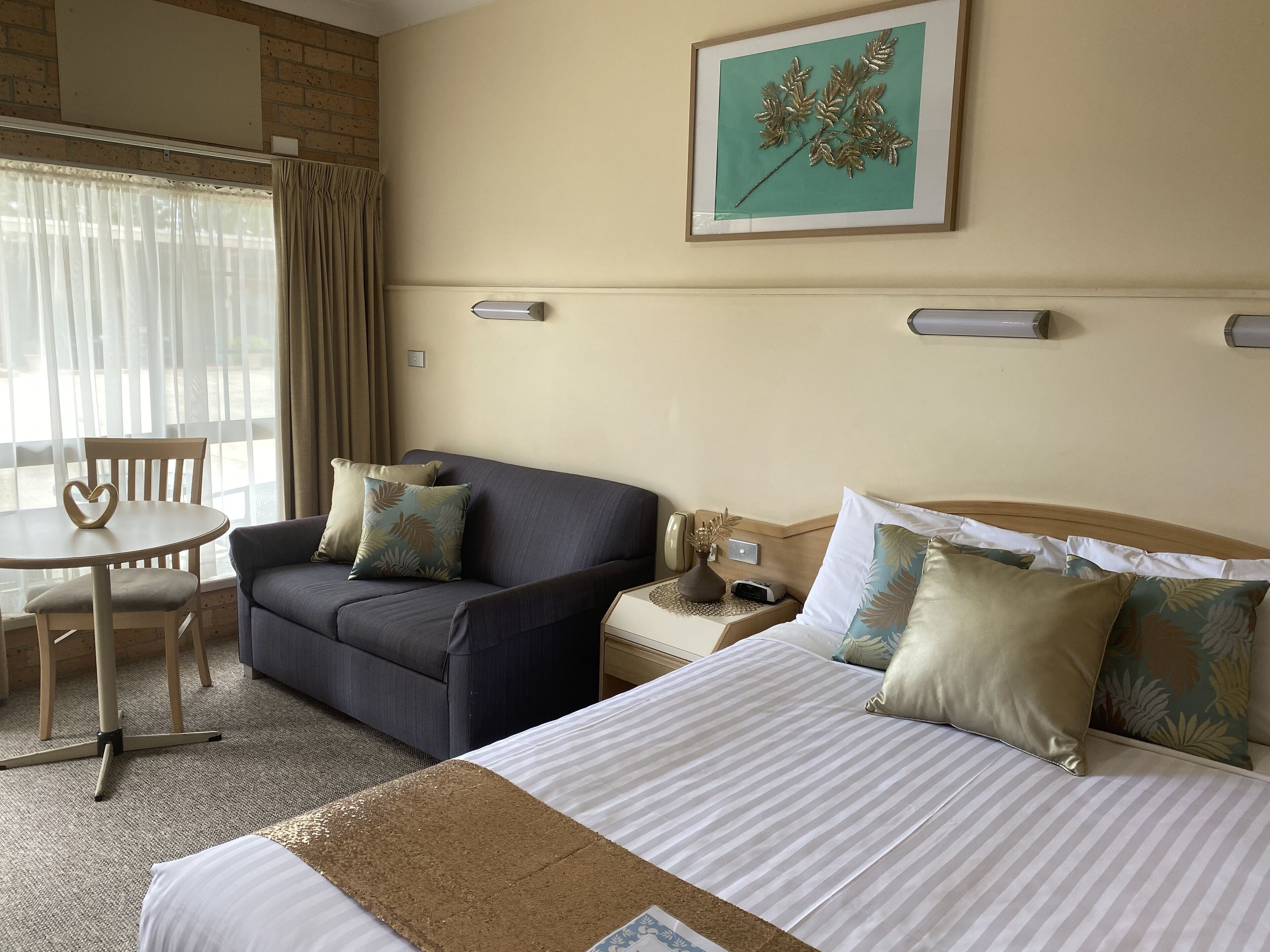 Habitación de hotel con cama, sofá, una mesita, y una silla. Se ve una ventana con cortinas, una lámpara de pared, y un cuadro en la pared.