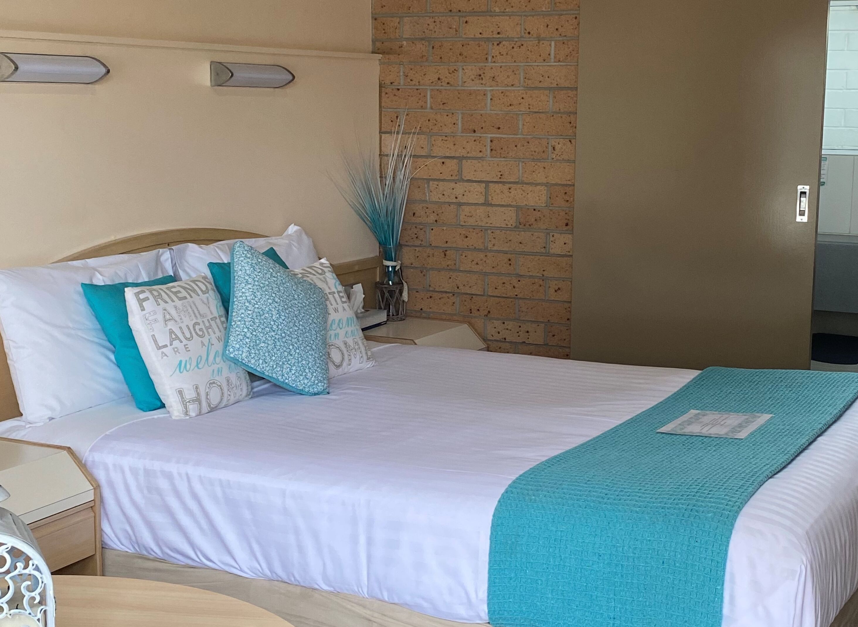 A neatly made bed with white bedding and teal accents, a brick wall behind, and a small bedside table with a lamp.
