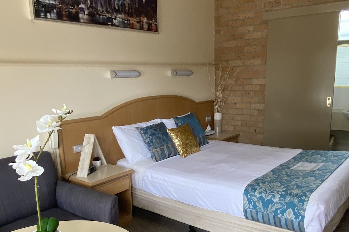 Quarto de hotel com cama, mesa de cabeceira, uma mesa com um vaso de flores, sofá e uma pintura de paisagem urbana na parede.