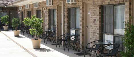 A row of outdoor tables and chairs with potted plants, a brick building with air conditioning units, and a clear sky.