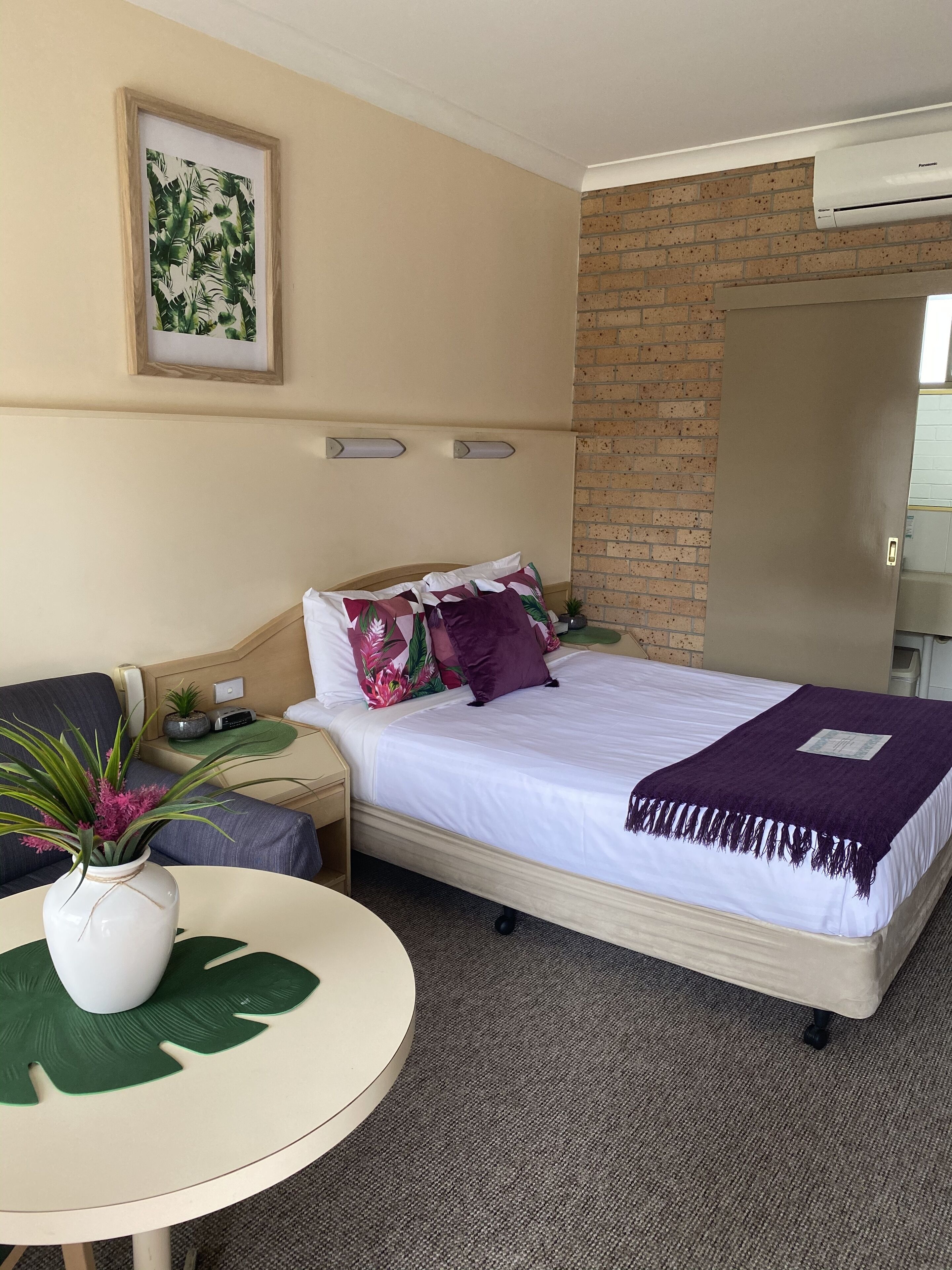 A hotel room with a bed, a bedside table with a plant, a small table with a plant, a picture on the wall, and a brick wall.