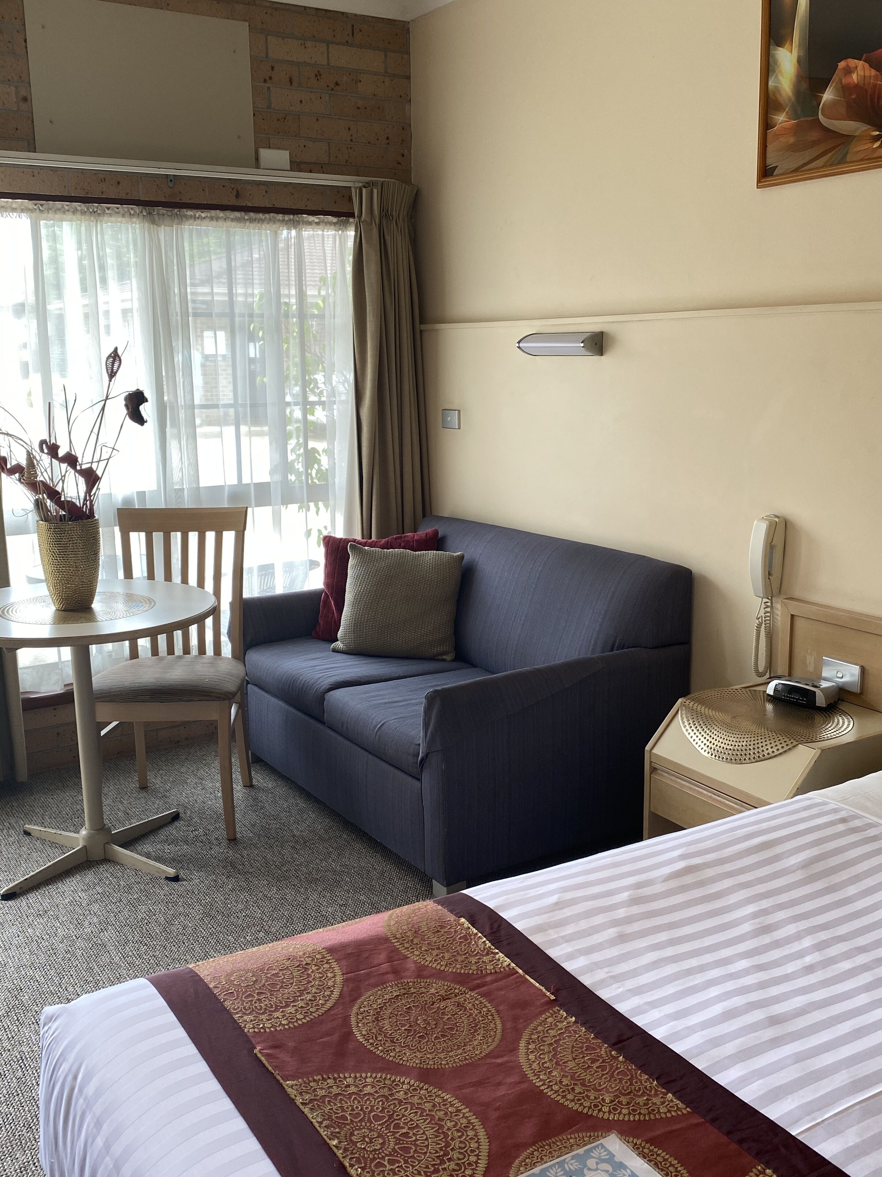 A hotel room with a bed, a sofa, a small table with a vase of dried flowers, a chair, and a nightstand with a phone and a lamp.