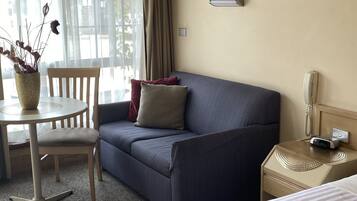 A hotel room with a bed, a sofa, a small table with a vase of dried flowers, a chair, and a nightstand with a phone and a lamp.