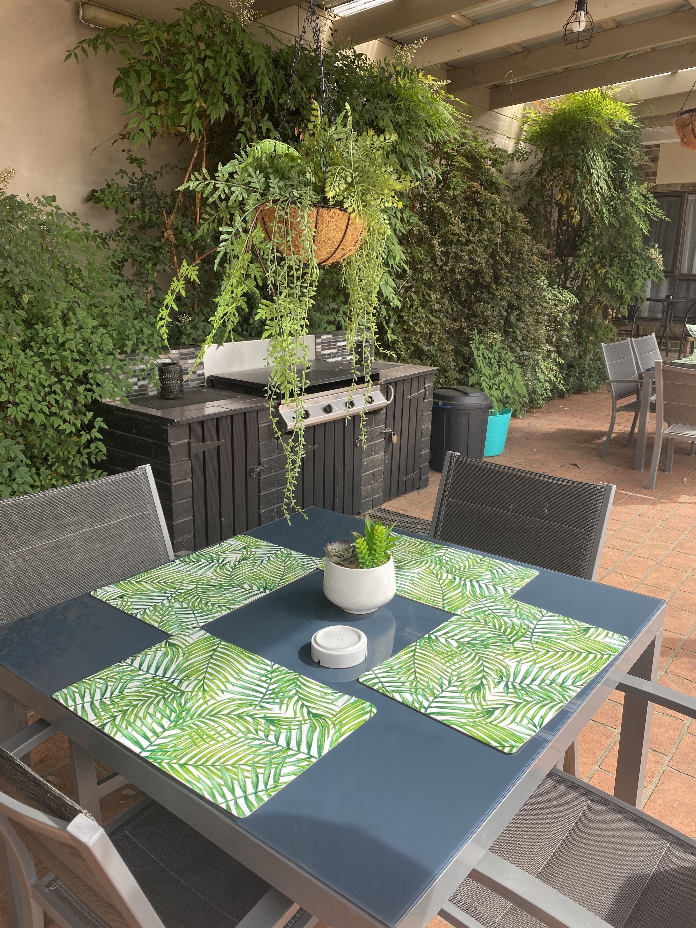 An outdoor patio with a table set for two, surrounded by lush greenery and a barbecue grill.
