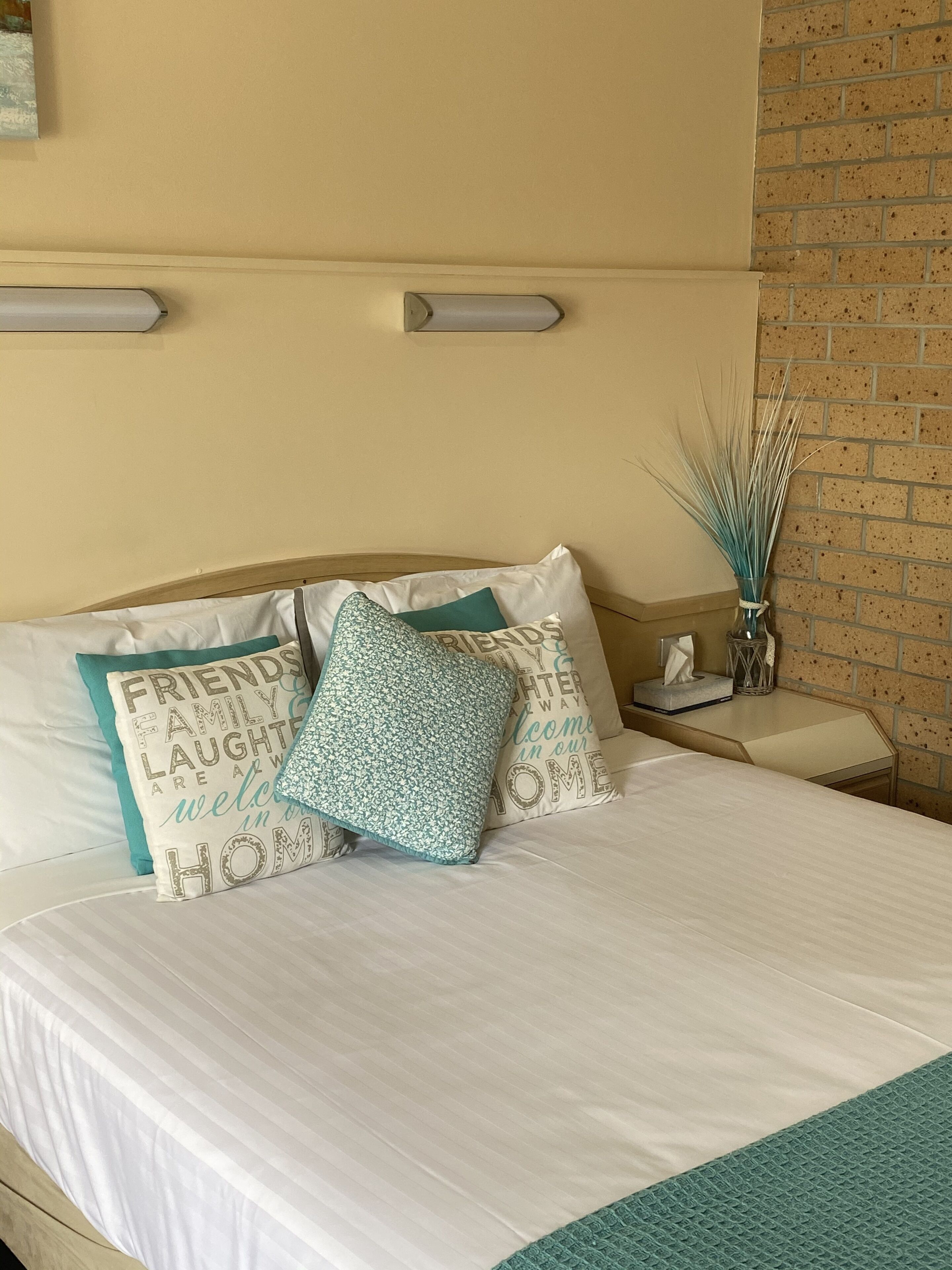 Un lit bien fait, avec une couette blanche et des touches de couleur turquoise, une table de chevet avec une boîte de mouchoirs et un vase décoratif contenant des branches.