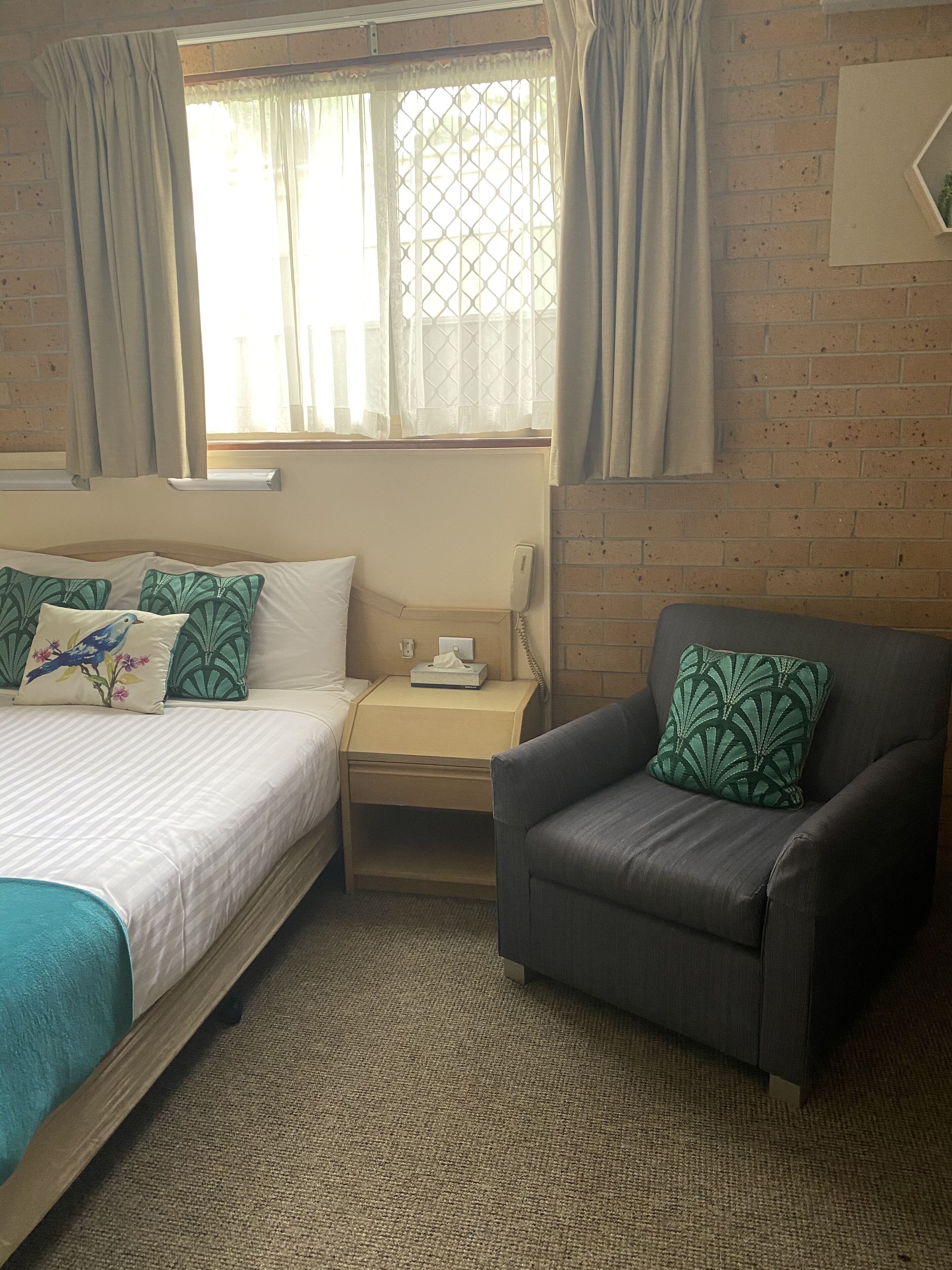Una habitación de hotel con cama, una silla, una mesita de noche con teléfono y ventana con cortinas.