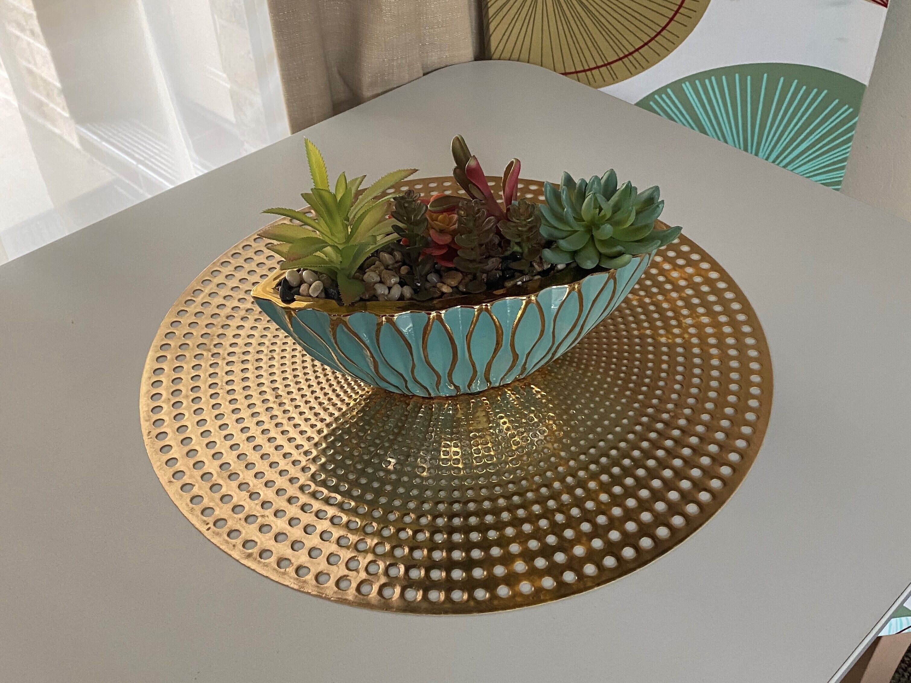 Un cuenco decorativo verde aguamarina y dorado con suculentas sobre una bandeja dorada perforada.
