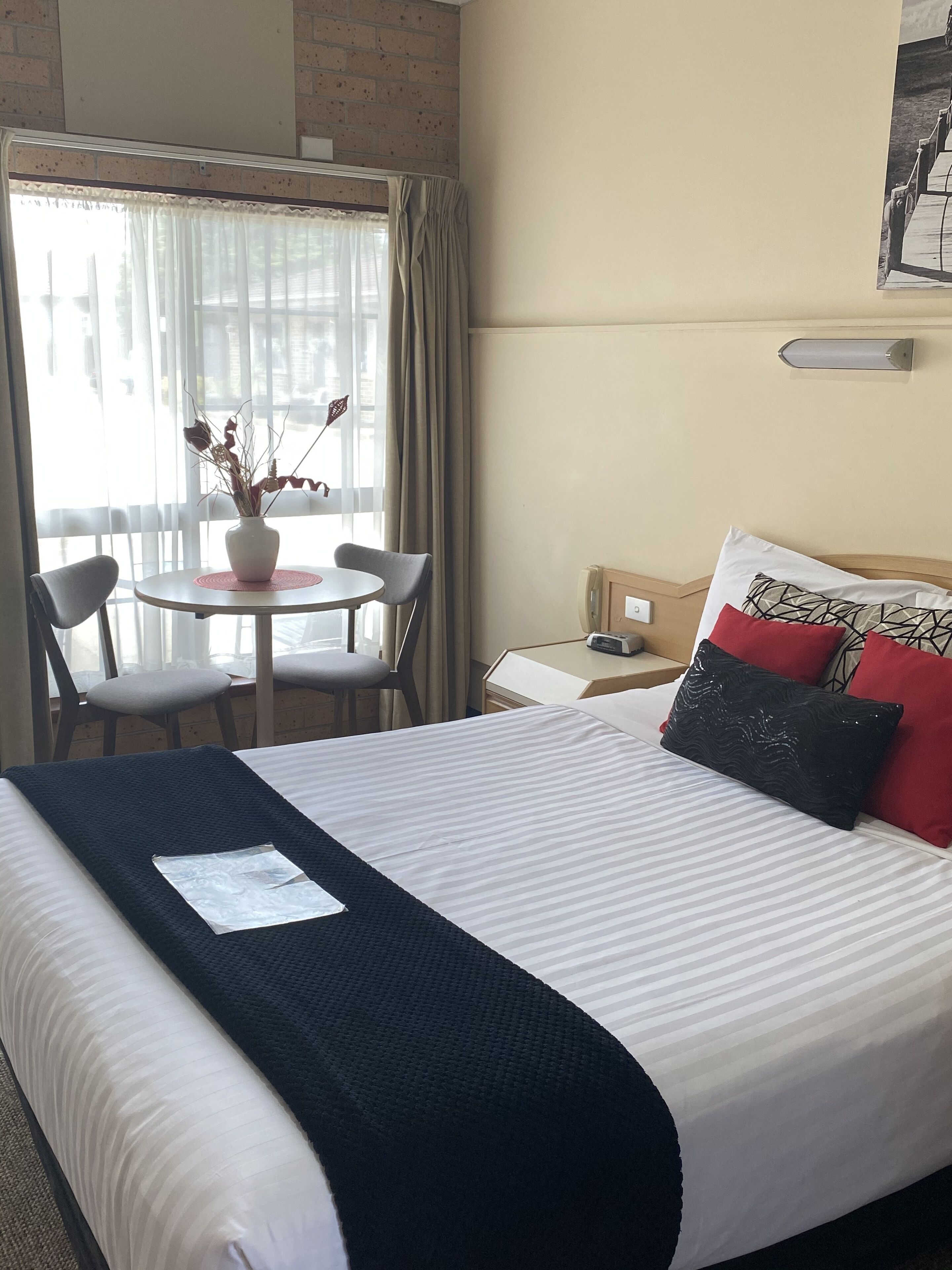 A hotel room with a bed, a small table with chairs, a window with curtains, and a wall-mounted light fixture.