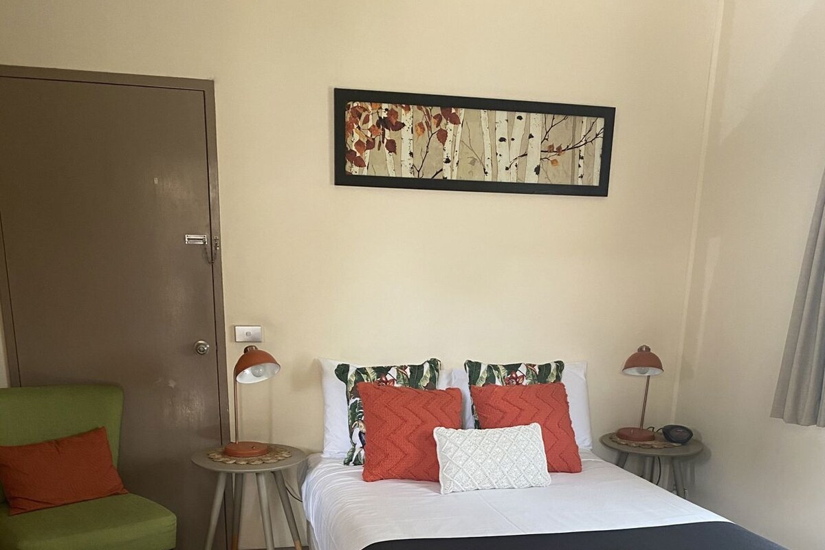 A hotel room with a bed, a chair, a small table, a lamp, and a painting on the wall.