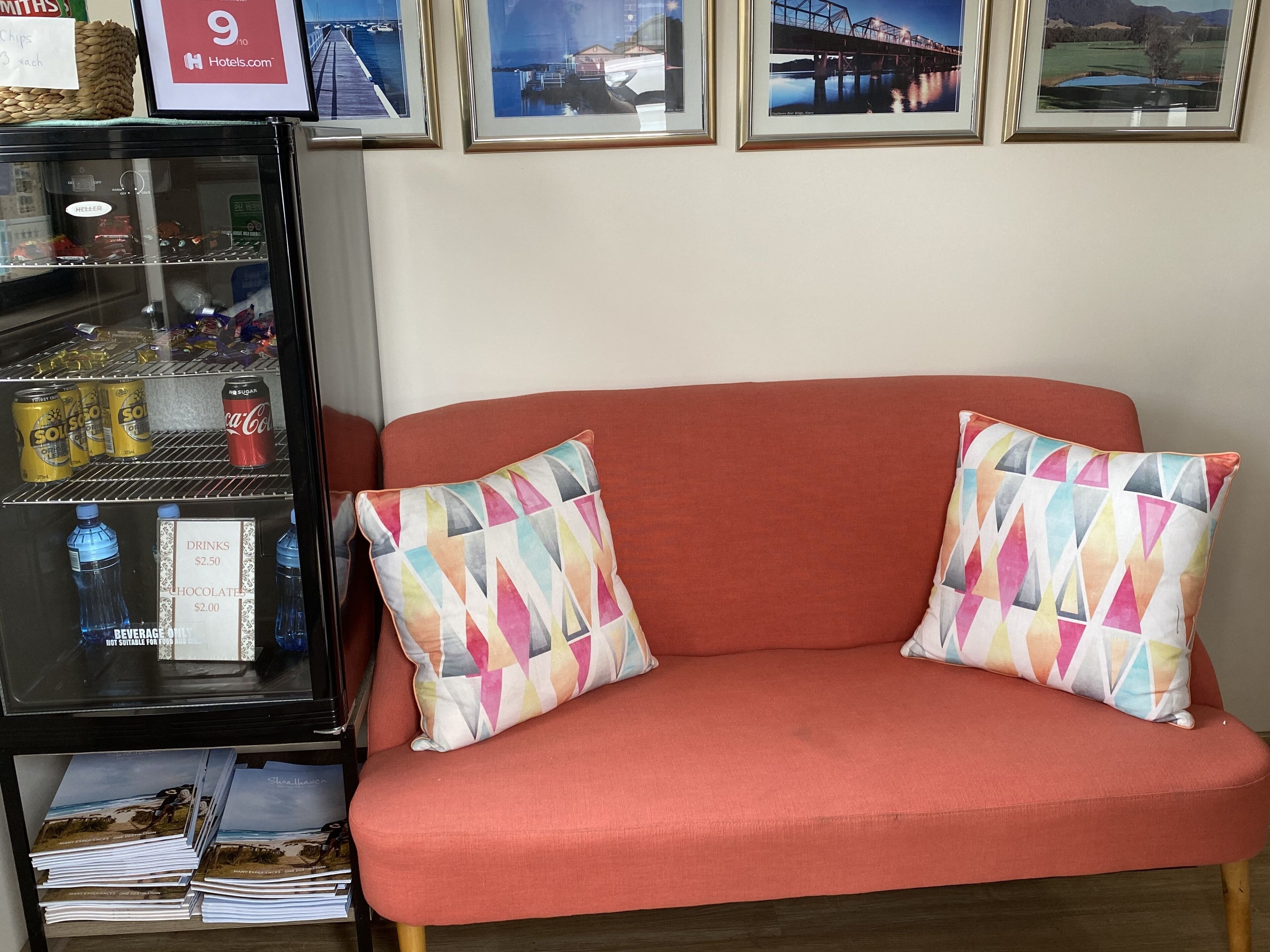 Une pièce avec un canapé rouge, deux coussins à motifs, un mini-bar contenant des collations et des boissons, et des tableaux encadrés accrochés au mur.
