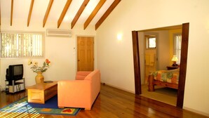 Una habitación con techo de madera, un sofá, una mesita, un televisor, una ventana con persianas y un espejo que refleja una cama.