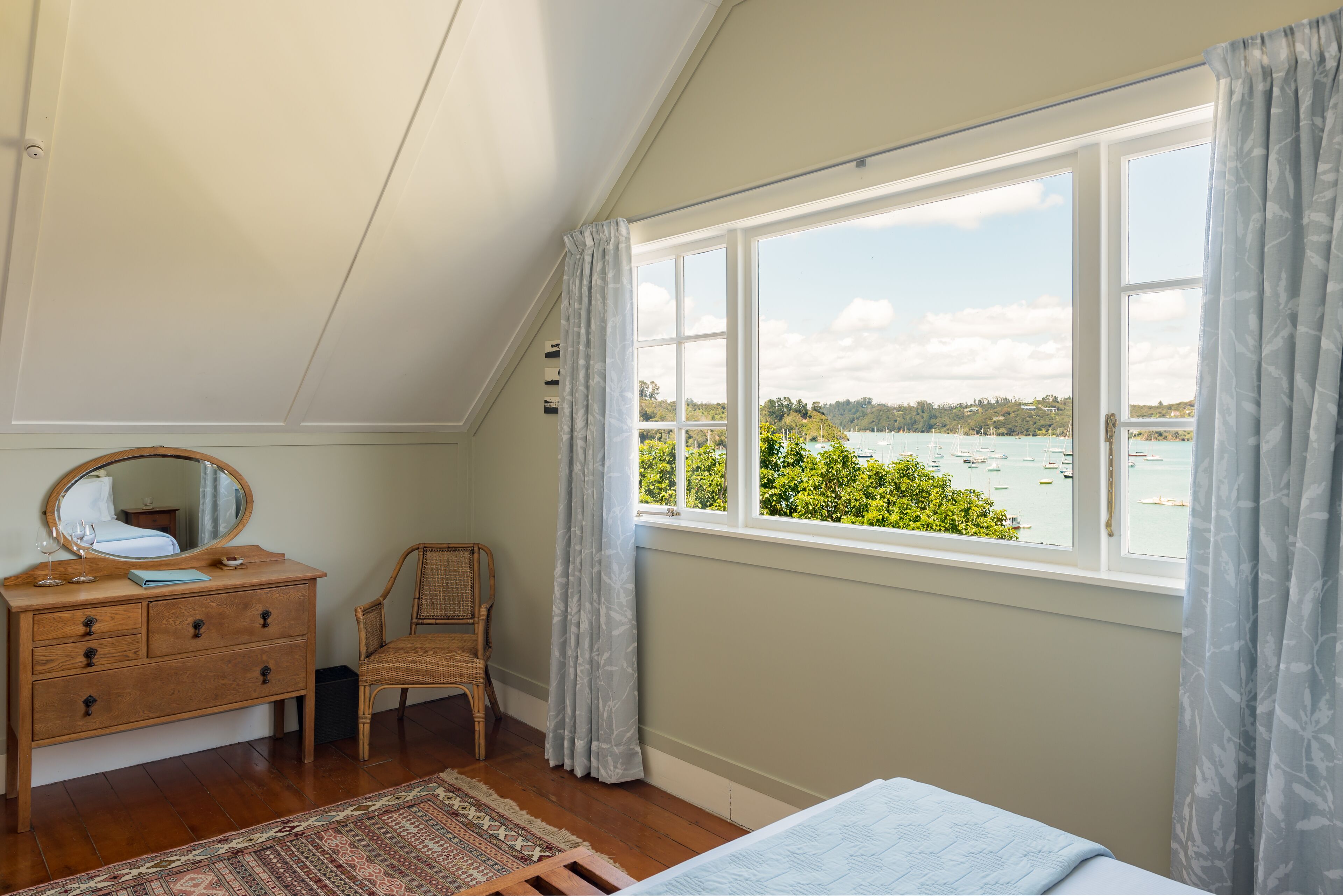 A bedroom with a wooden dresser, a chair, a bed, and a large window offering a view of a body of water and greenery.