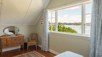 A bedroom with a wooden dresser, a chair, a bed, and a large window offering a view of a body of water and greenery.