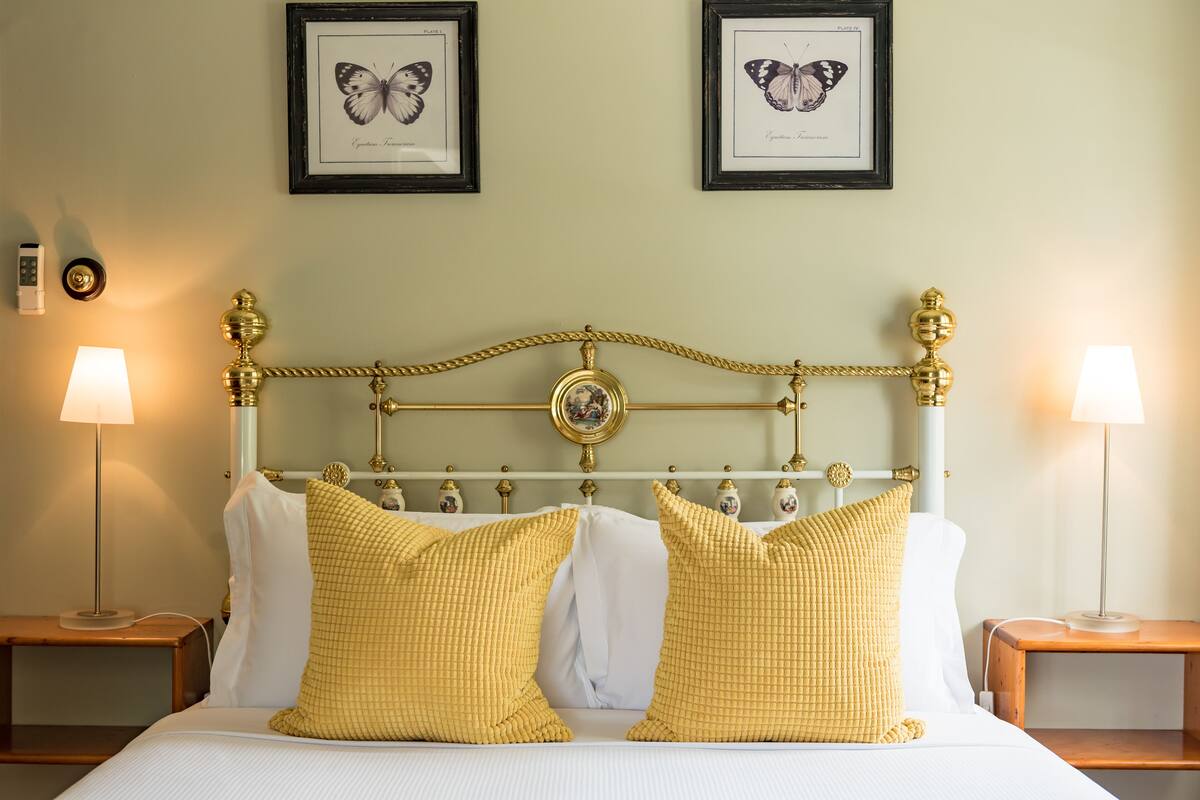 Un lit bien fait, agrémenté d’oreillers blancs et jaunes, d’une tête de lit encadrée d’or et de deux tableaux représentant des papillons accrochés au mur.
