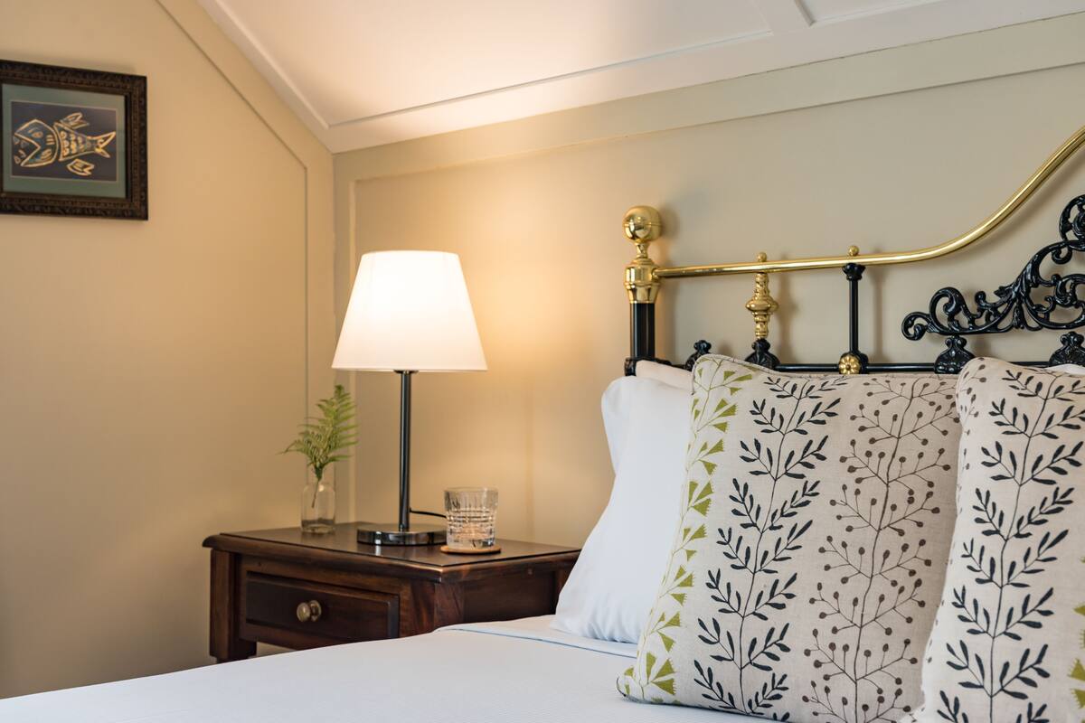Un lit bien fait, recouvert d’une couette blanche et agrémenté d’un coussin décoratif orné d’un motif de feuilles. Sur une table de chevet, on trouve une lampe, un verre d’eau et un petit vase avec une plante.