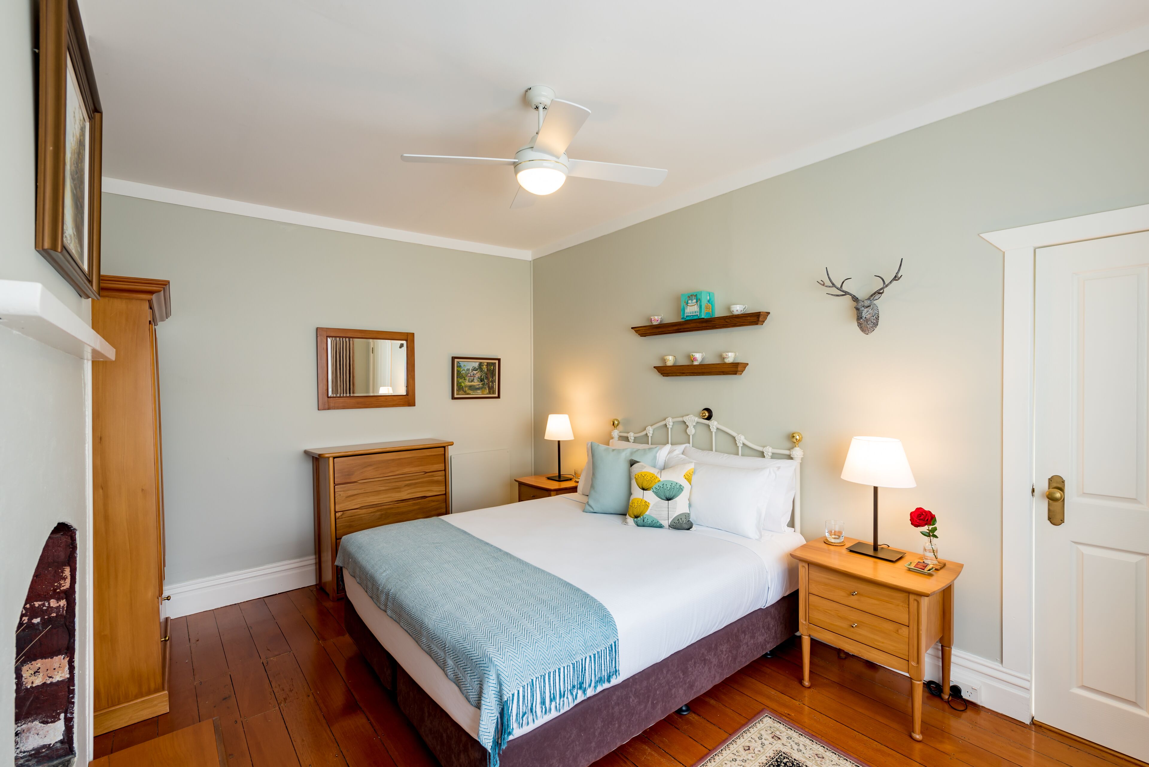 Un dormitorio con cama, mesitas de noche, ventilador de techo, suelo de madera, puerta y estante montado en la pared con objetos decorativos.