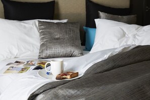 A neatly made bed with a white and gray duvet, a blue pillow, and a white pillow. A slice of toast and a white mug are on the bed.