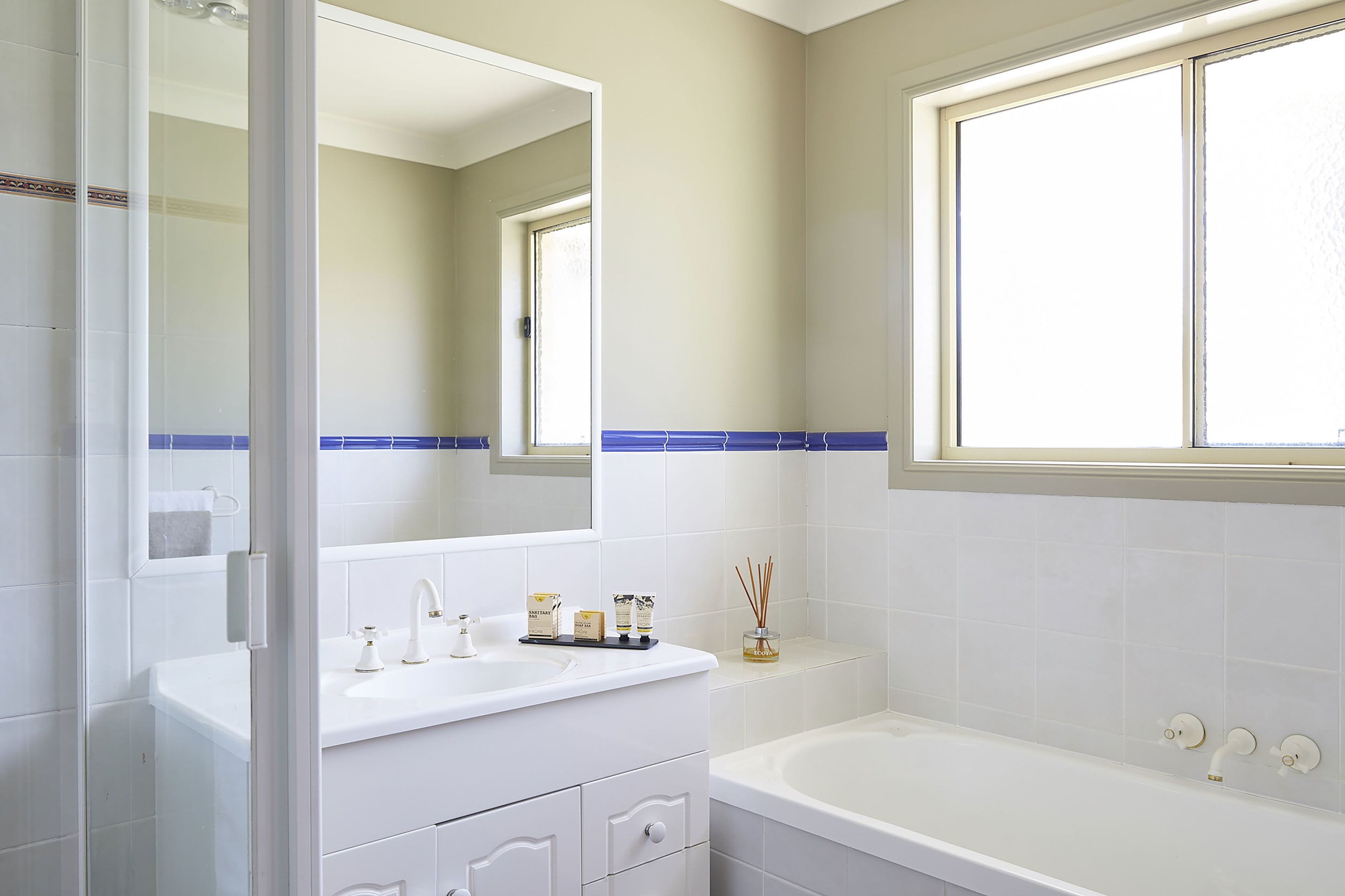 A bathroom with a white sink, mirror, and bathtub. There is a window above the bathtub and a shelf with toiletries.