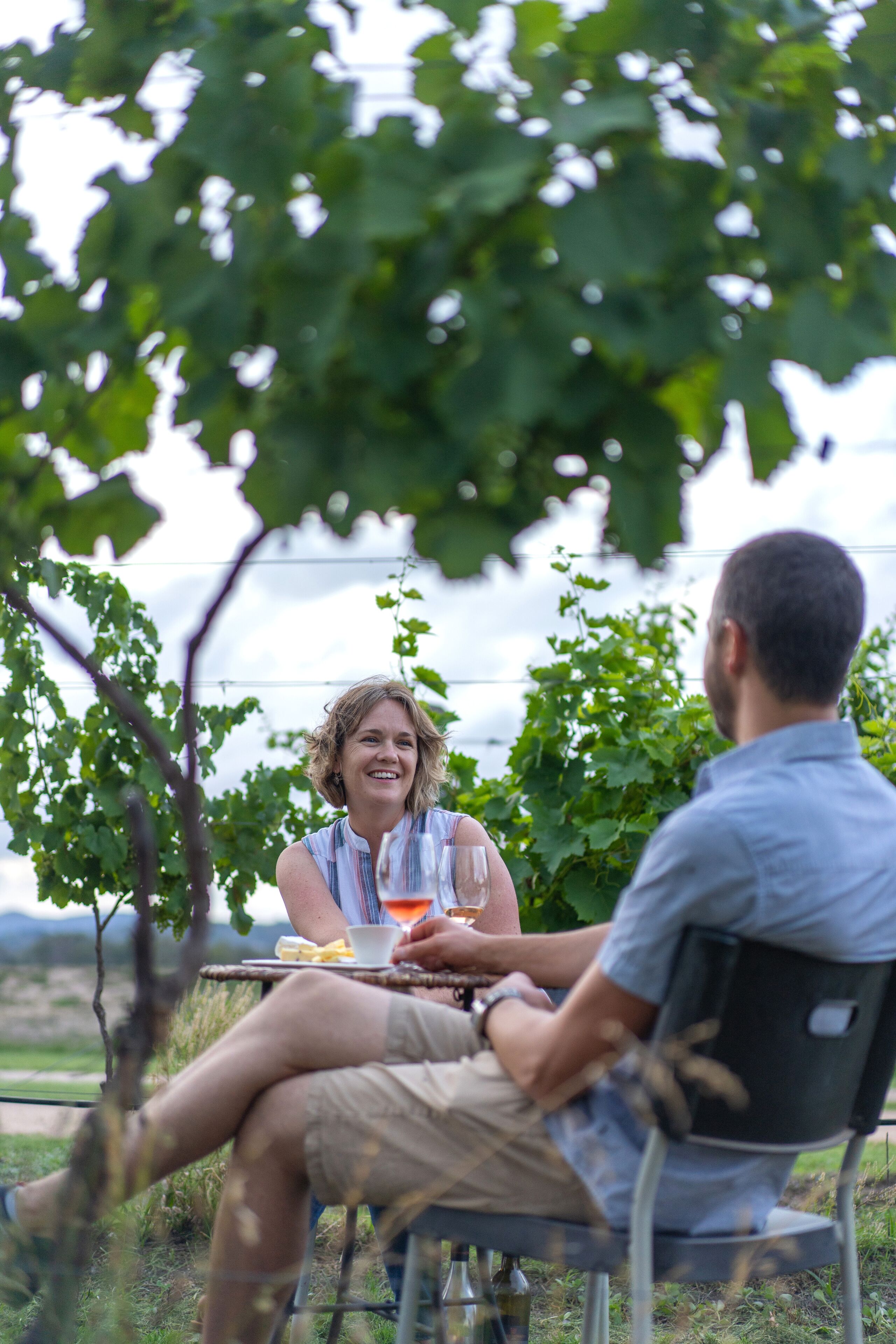 Zwei Personen sitzen an einem Tisch im Freien, mit Getränk und Speise, umgeben von viel Grün und blauem Himmel.