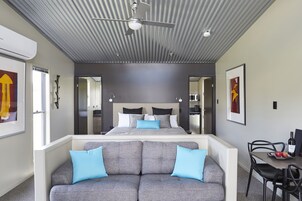 A modern bedroom with a large bed, a ceiling fan, a dining area with a table and chairs, and a sofa with blue cushions.
