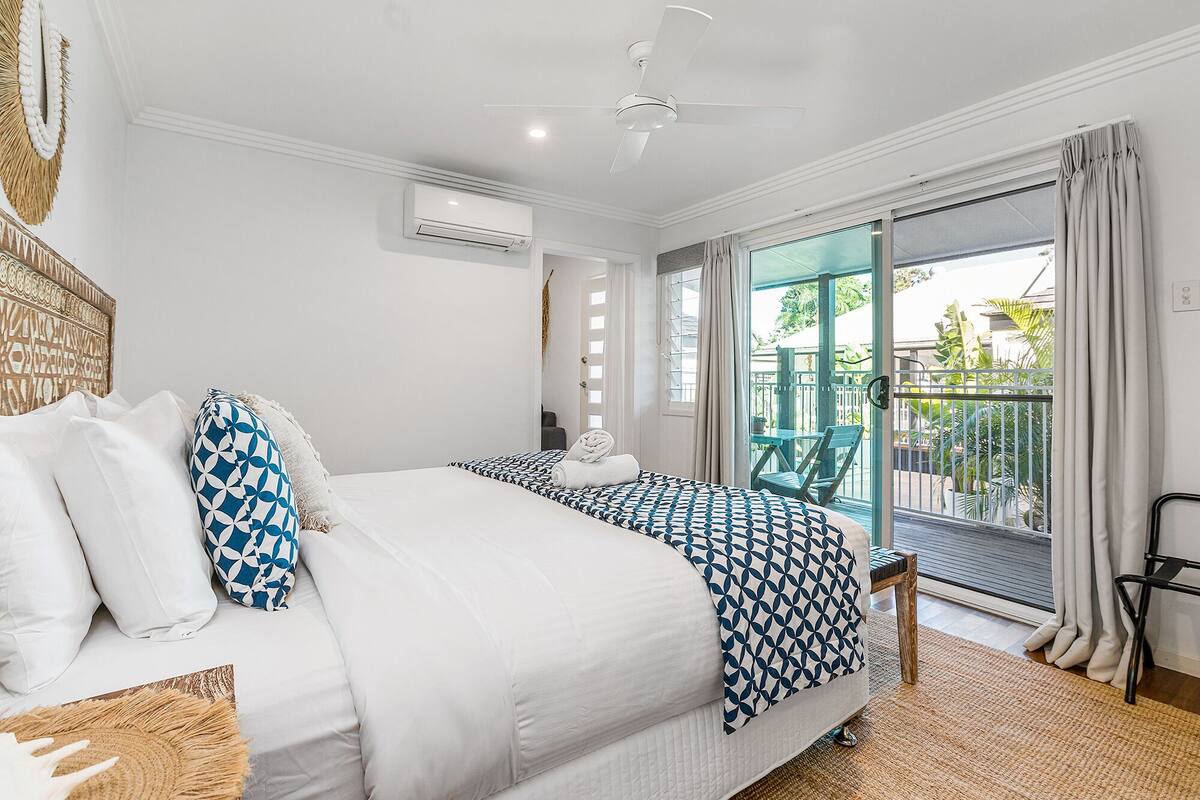 A bedroom with a large bed, a ceiling fan, an air conditioning unit, and a view of the outside balcony.