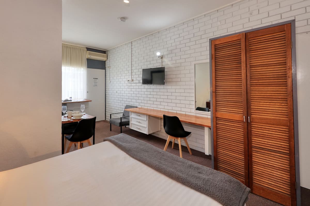 A hotel room with a bed, a desk with a chair, a television mounted on the wall, and a wooden wardrobe.