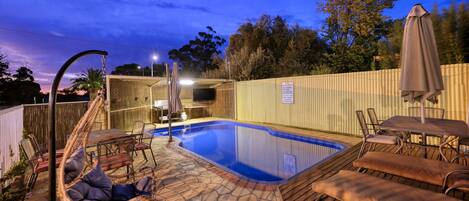 A backyard with a swimming pool, lounge chairs, a TV, and a wooden fence.
